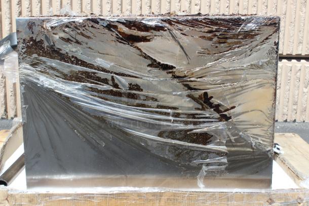 Large rectangular metal sheet wrapped in protective plastic, resting on a wooden pallet. Slight oxidation visible.