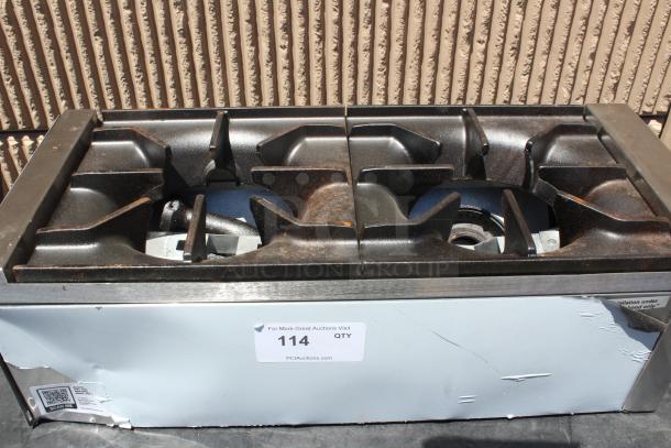 Commercial gas stovetop with four burners, metal frame, noticeable rust, and dents. Auction tag reads "114 QTY PCAuctions.com".