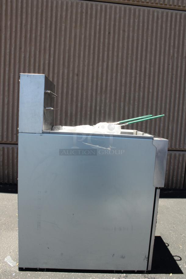 Commercial stainless steel fryer, side view, showing minor surface scratches and wrapped accessories on top.