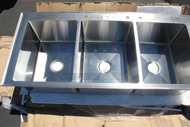 Stainless steel triple-basin sink in new condition, with pre-drilled faucet holes, resting on cardboard packaging.