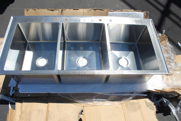 Triple-basin stainless steel kitchen sink, unused, with dual faucet holes and visible model tag labeled "134" on the rim.