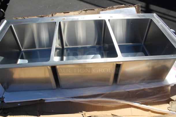 Stainless steel triple bowl kitchen sink, unused, with installation holes, set on cardboard packaging.