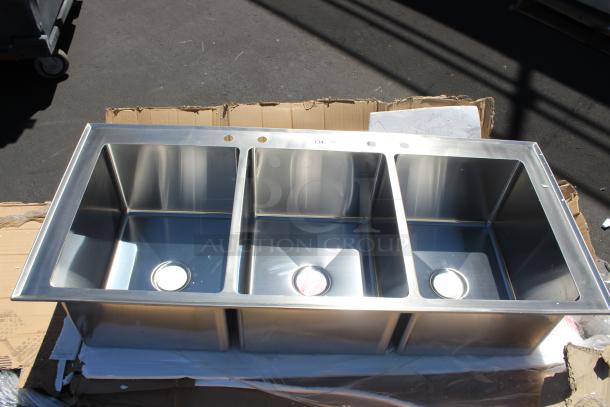 Stainless steel triple basin sink, new condition, with pre-drilled faucet holes and drain covers visible. Label reads "134."