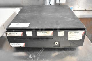 Black metal cash drawer with key lock, visible service labels, moderate surface wear, size similar to standard office drawer.