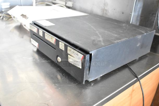 Black metal cash drawer with front lock, visible labels, and slight surface scratches, in used condition.