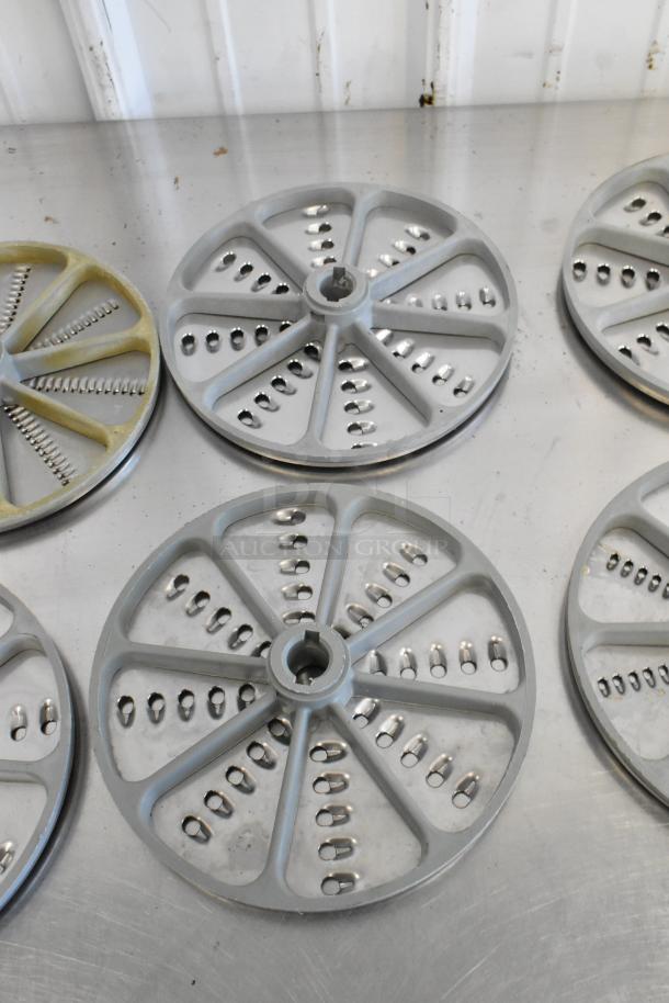 Ten various food processor blades, metal and plastic, used condition, on a stainless steel surface. Multiple shredding designs.