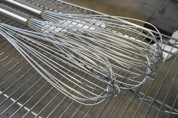 Three new metal mixing whisks with long handles, scratch and dent condition, displayed on a metal rack.