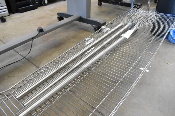 Three brand new metal mixing whisks on a rack, featuring long handles and slight scratches, with tags attached.