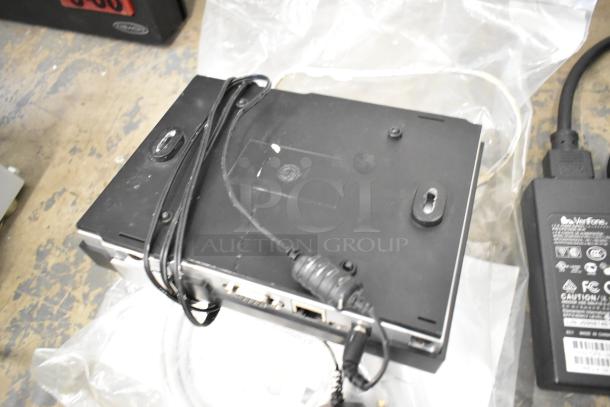 "Lot of items including a black Verifone credit card reader with cables, in used condition, placed on a textured surface."