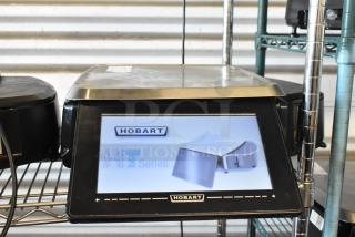 Hobart HT series digital scale, touchscreen display, metal casing, in good condition on a metal shelf.