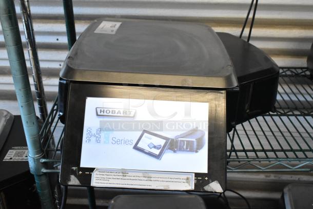 Hobart HT Series scale, black, with digital display, visible branding and instructions, in used condition on metal shelf.