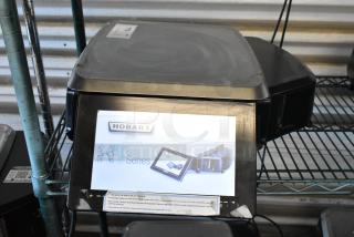 Hobart HT Series scale, black, with digital display, visible branding and instructions, in used condition on metal shelf.