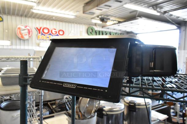 Hobart commercial kitchen touchscreen scale, black, mounted on a metal rack in a warehouse setting.