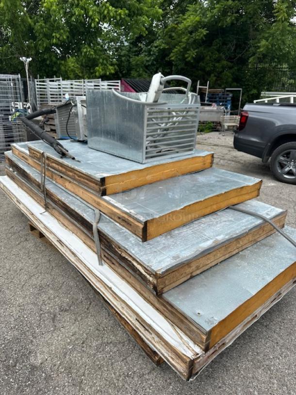 Stack of insulated metal panels with exposed wood edges on a pallet, accompanied by a metal basket with tools. Panels show slight wear.