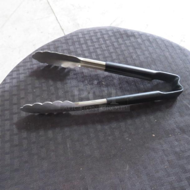 Metal kitchen tongs with black handles, placed on a patterned surface, in good condition.
