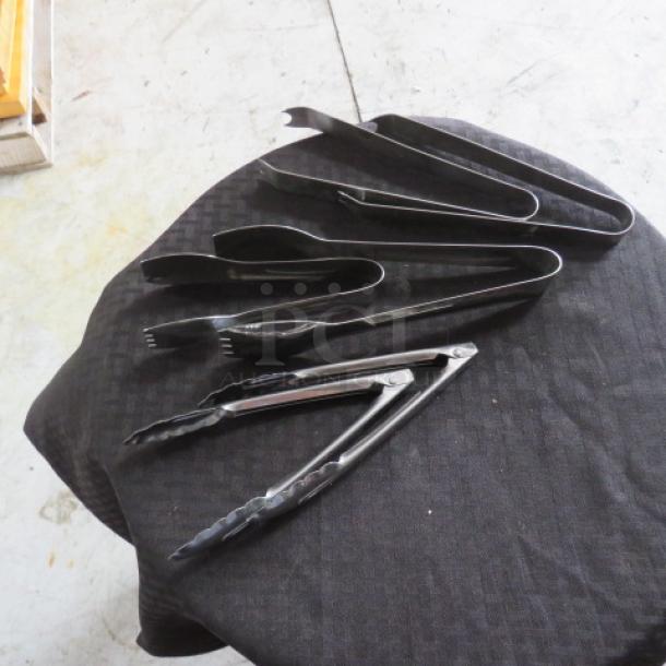 Set of five stainless steel kitchen tongs on a black cloth, varying in size and design, in good condition.