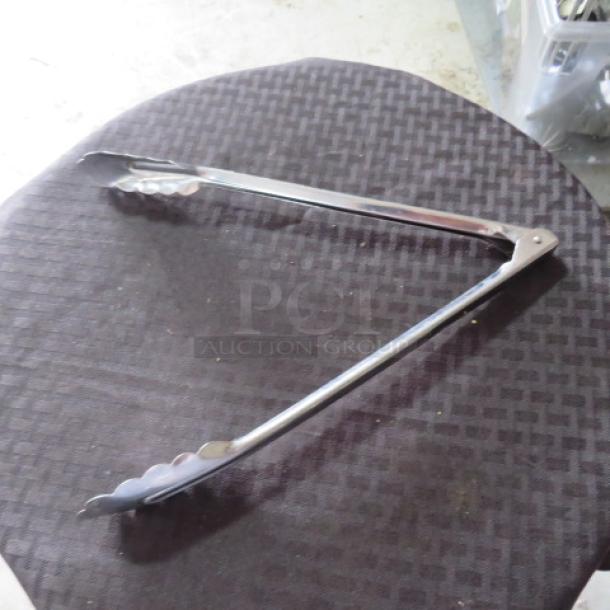 Stainless steel kitchen tongs with scalloped edges, resting on a dark surface. In good condition, showing minor wear.