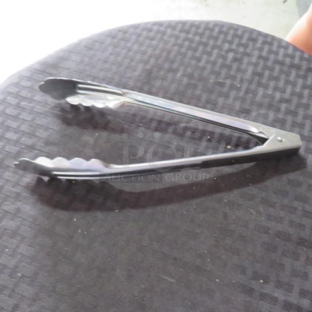 Metal kitchen tongs with scalloped edges, silver finish, in good condition, laid on a textured black surface.
