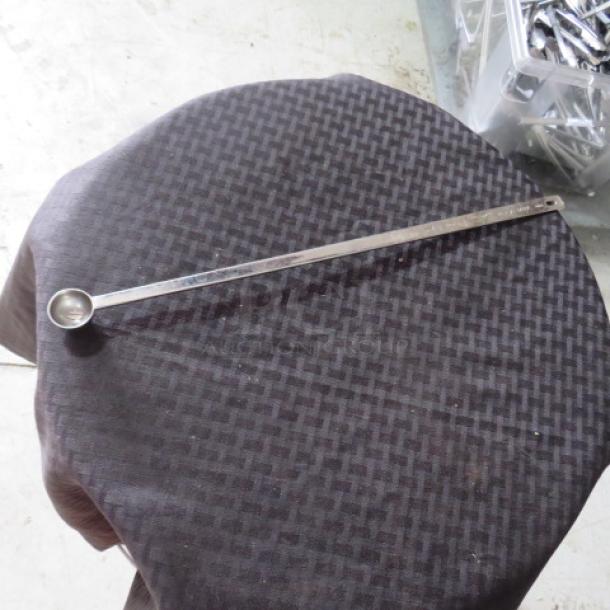 Long metallic spoon with small, deep bowl, lying on a checkered tablecloth. Appears in good condition, no visible markings.