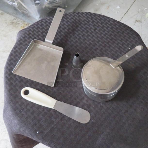 Set of stainless steel kitchen tools on table: a flat spatula, dough scraper, icing nozzle, and lidded pot, in used condition.