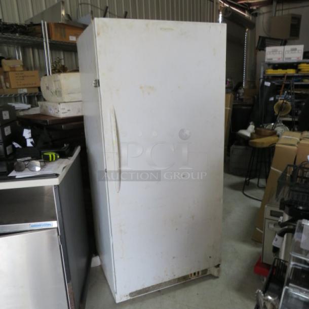 White upright freezer in used condition, single door, metal handle, slight surface marks, no visible brand or model.
