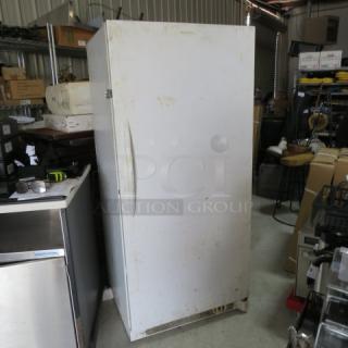 White upright freezer in used condition, single door, metal handle, slight surface marks, no visible brand or model.