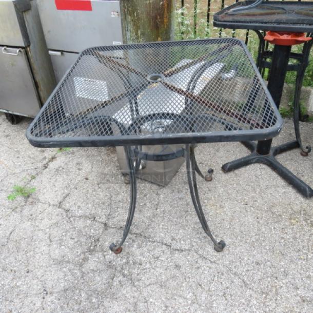 Black metal patio table with mesh top, square design, curved legs, lightly rusted, suitable for outdoor use.