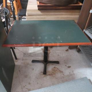 Rectangular table with green laminate top, wood-trimmed edges, and black metal pedestal base. Visible surface wear.