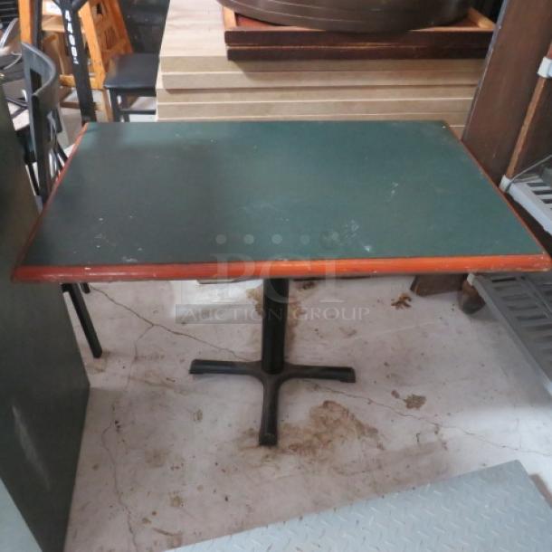 Rectangular dining table with green laminate top and wooden trim, showing minor wear. Black metal pedestal base.