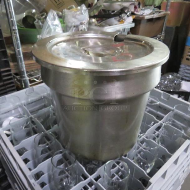 Large stainless steel commercial soup kettle with lid, situated in a dish rack. Some surface wear, indicating use.