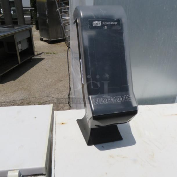 Tork Xpressnap napkin dispenser, black, upright design, outdoor setting, "Napkins" embossed on front, good condition.