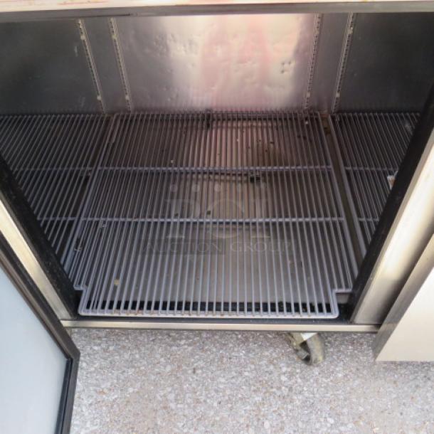 Commercial stainless steel refrigerator interior with shelving, open door, moderate wear, visible floor casters.