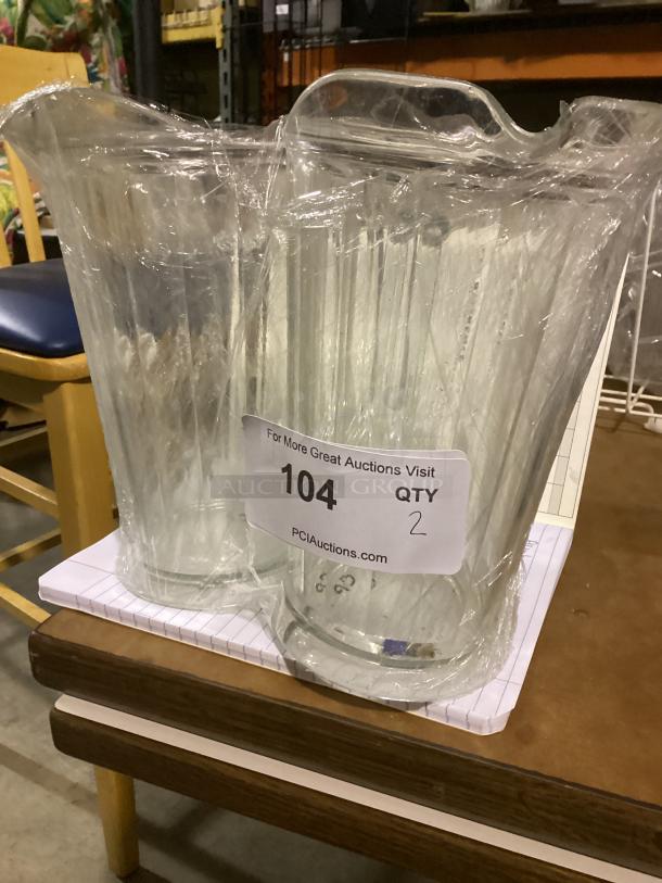 Two clear plastic juice pitchers, 9-inch height, wrapped in plastic, label indicates quantity 2, item number 104.