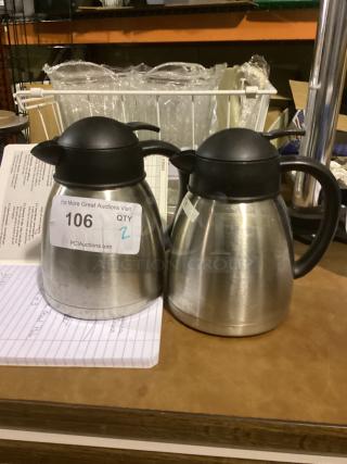 Pair of stainless steel coffee pots with black lids, labeled "106 QTY 2" at auction. Good condition.