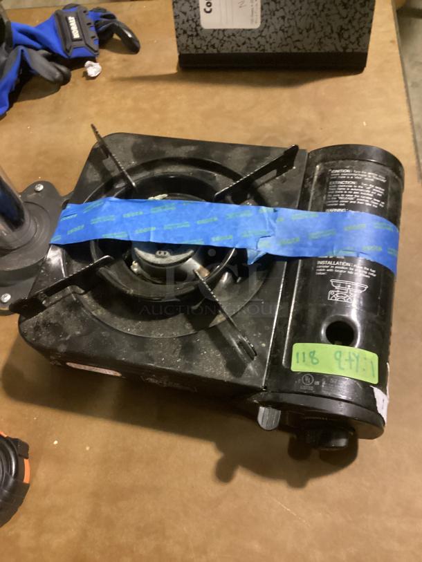 Portable butane stove with single burner, used condition, safety labels visible, secured with blue tape, model 13-1/8.