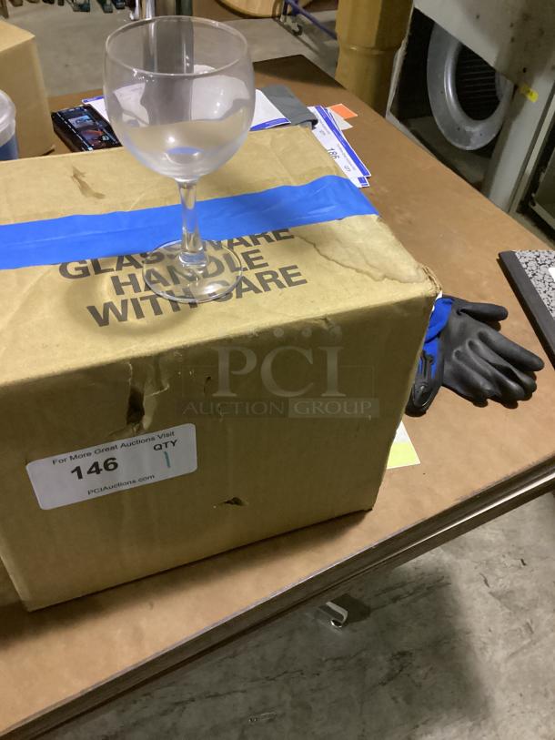 Red wine glass, standing on a labeled cardboard box with "Glassware Handle With Care." Box shows item quantity and auction details.