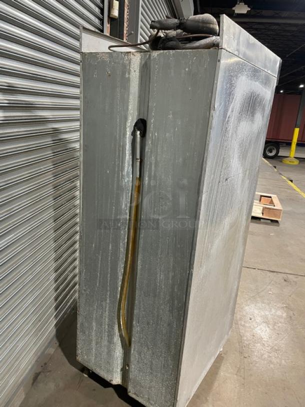 Commercial stainless steel refrigerator, used condition, visible wear, cooling pipes exposed, industrial setting.