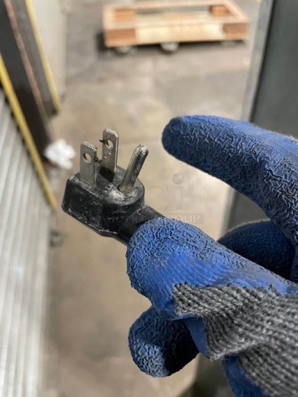 Worn electrical plug with three prongs, held by a gloved hand. Some damage visible; functional status uncertain.