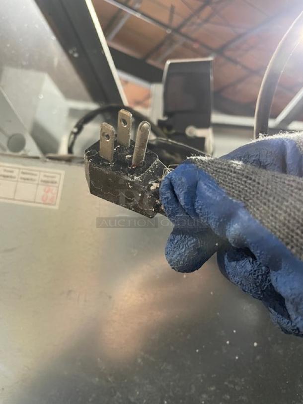 Three-prong electrical plug held in gloved hand, showing wear. Visible labels on background equipment indicate specifications.