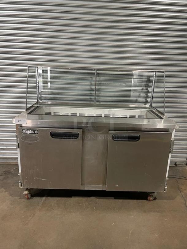 Stainless steel food prep counter with glass sneeze guard, two doors, caster wheels; brand "Lando", moderate wear visible.
