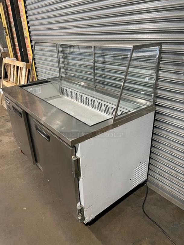 Commercial stainless steel refrigerated prep table with glass sneeze guard. Features two-door storage, moderate wear. Ideal for restaurant use.