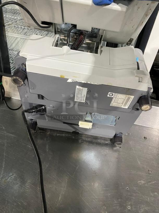 Office copier machine, grey, used condition. Visible power cables and labels with technical specifications on the side.
