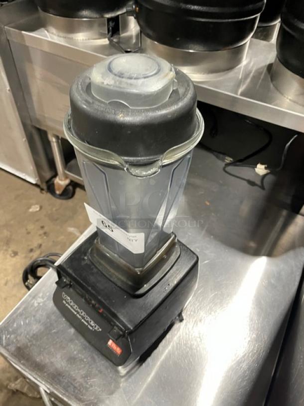 Commercial blender on metal counter, used condition, black base, clear container, visible power switch, price tag labeled "65".