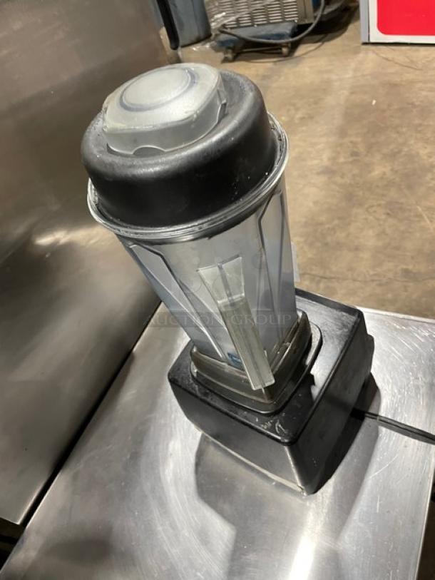 Industrial blender with a transparent, ribbed container and a black base. Appears in used condition with wear visible.