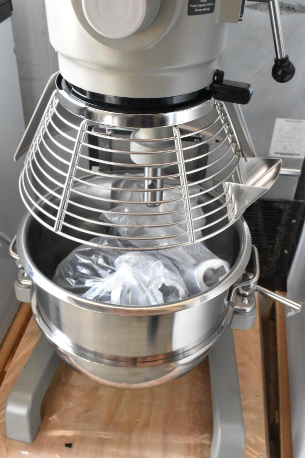 Commercial stand mixer with stainless steel bowl, safety guard, and attachments in plastic wrap. Appears in new condition.