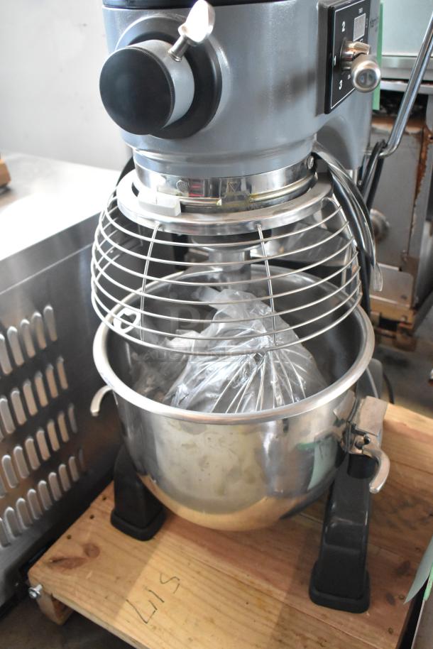 Commercial stand mixer with metal bowl, safety guard, and variable speed control. Placed on wooden platform. Good condition.