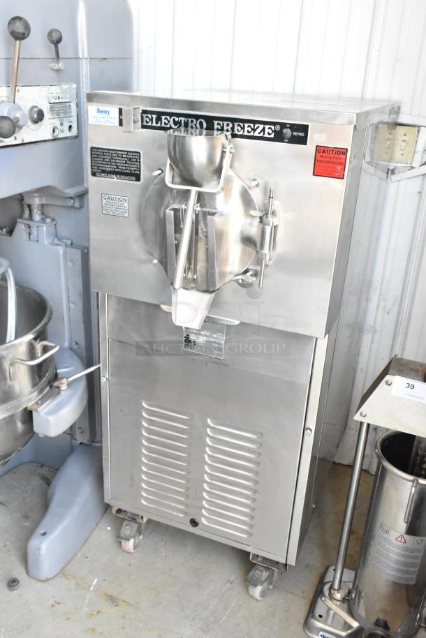 Electro Freeze ice cream machine, stainless steel, model 30T-RMT, used, caution labels visible, on casters for mobility.