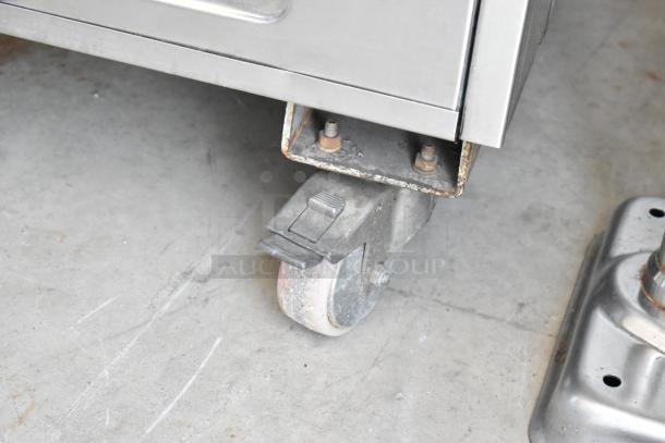 Metal industrial caster with a locking mechanism on a welded steel base. Slight rust on bolts, indicating some wear.