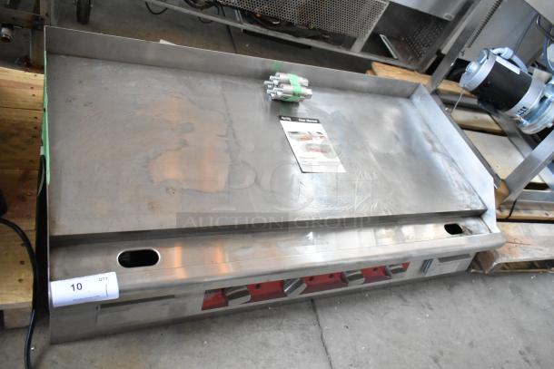 Commercial stainless steel griddle with control knobs, user manual, and metal parts on top. Some surface wear visible.