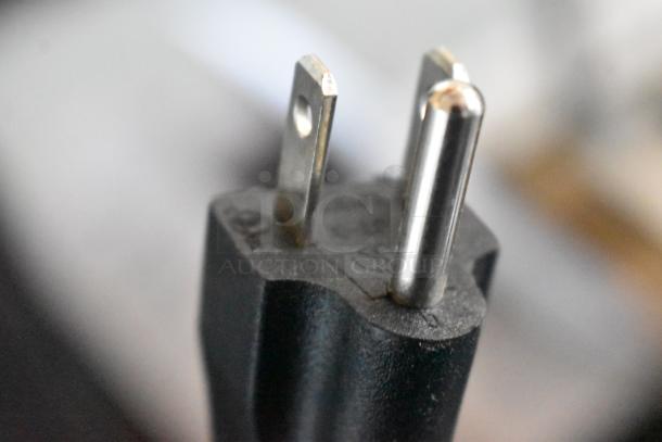 Close-up of a three-prong electric plug. Black casing, metallic prongs. Good condition. Indistinct markings on the surface.